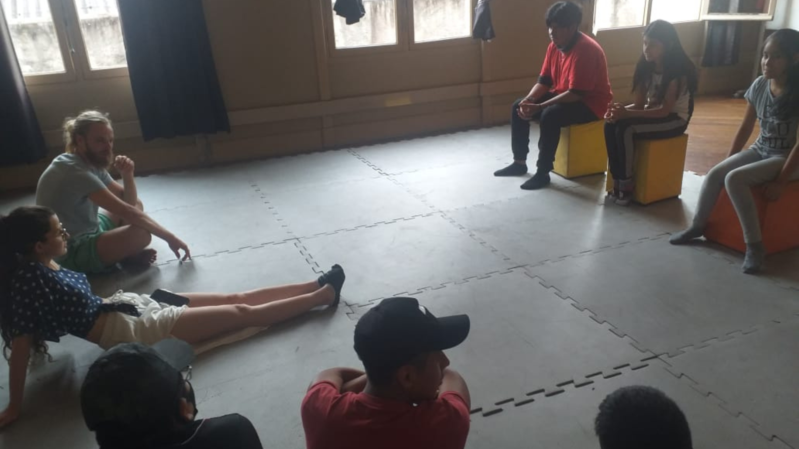 Estudantes-bolsistas da SP Escola de Teatro e crianças e adolescentes bolivianos da região da sede Brás estão sentados em círculo realizando atividades artísticas