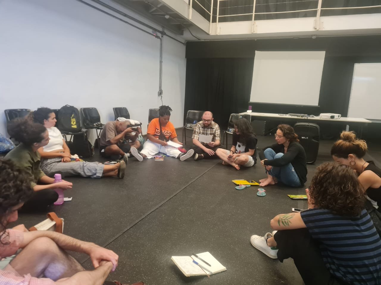 Fotografia colorida de pessoas sentadas no chão, em roda, durante aula na SP Escola de Teatro. 