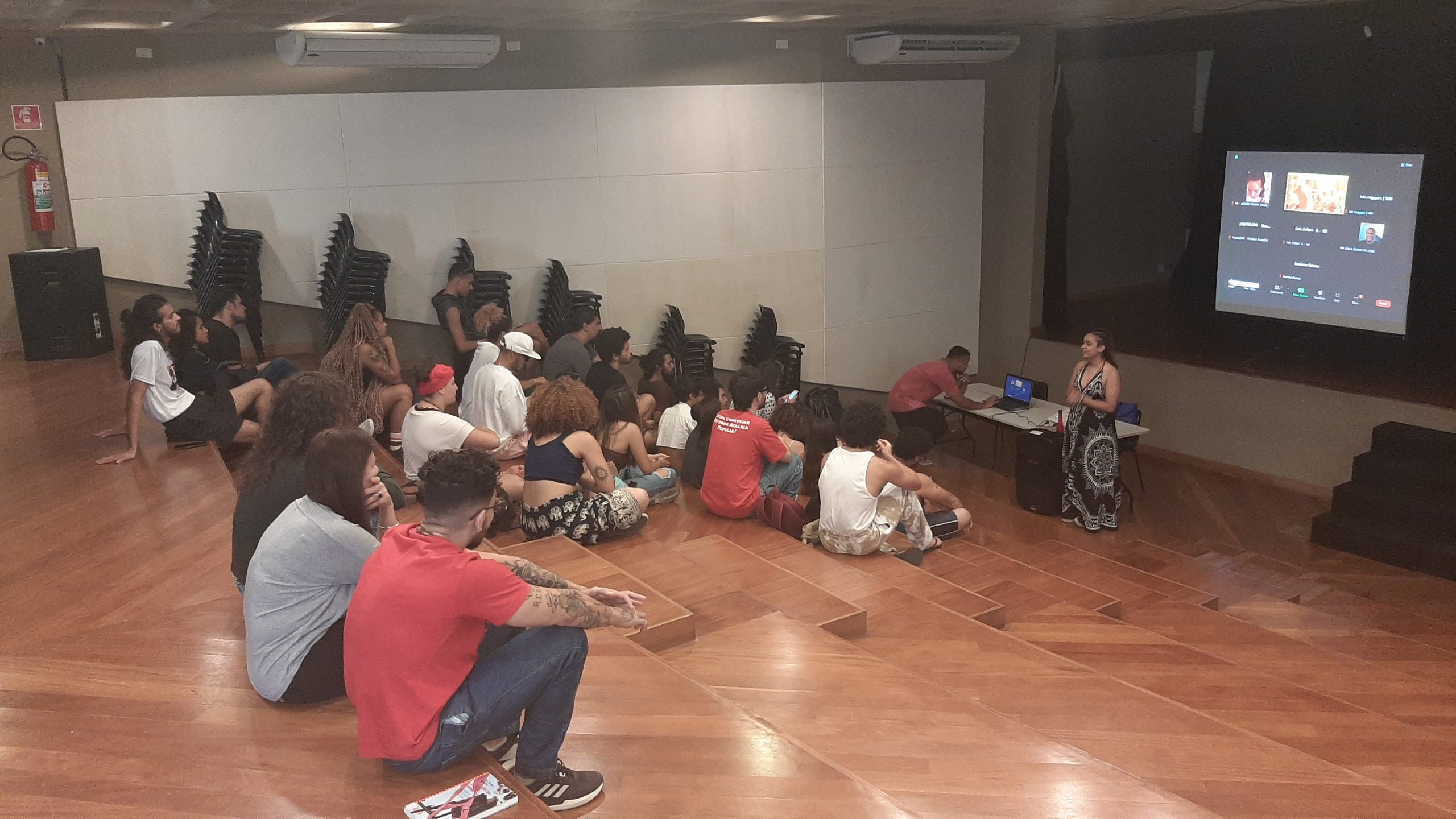 Foto colorida mostrando os estudantes da SP Escola de Teatro sentados no tablado do auditório da sede Brás