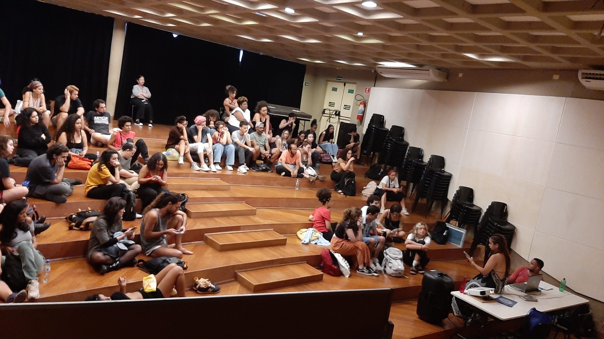 Foto colorida mostrando os estudantes da SP Escola de Teatro sentados no tablado do auditório da sede Brás
