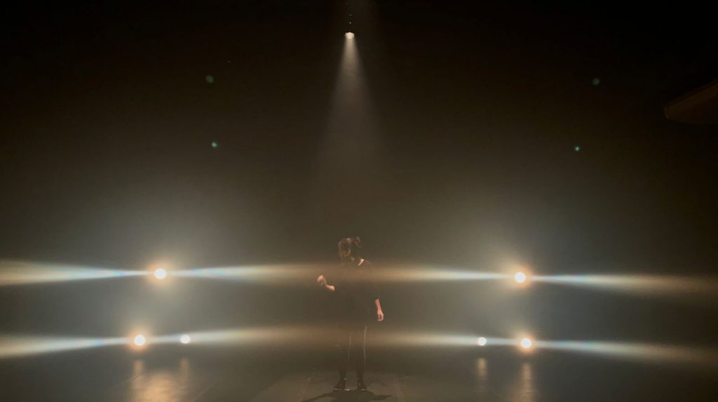 Foto com uma pessoa no palco sob a luz amarela formando duas linhas horizontais