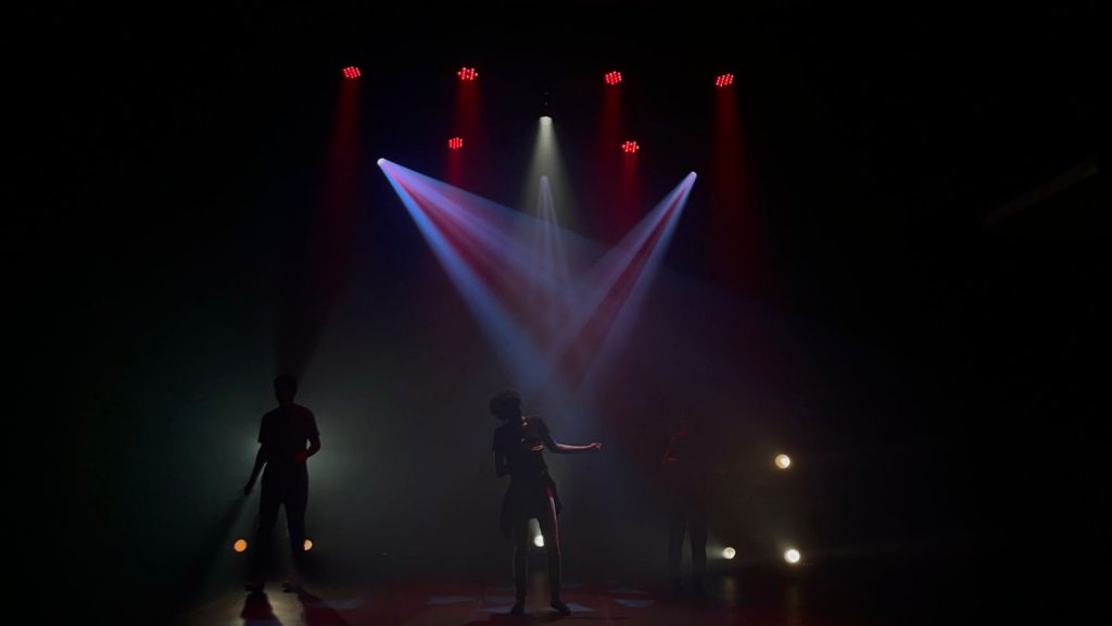 Foto escura mostrando três pessoas no palco sob a iluminação da combinação das luzes vermelha e azul