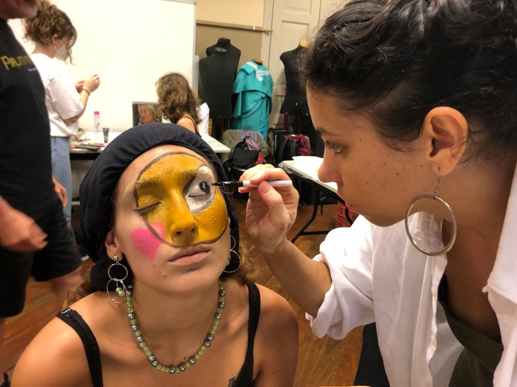 Foto colorida mostrando uma estudante fazendo maquiagem em outra estudante. A maquiagem tem a cor amarela, com contorno preto e detalhes em branco e rosa.