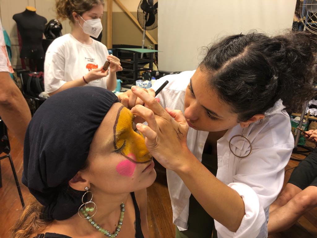 Foto colorida mostrando uma estudante fazendo maquiagem em outra estudante. A maquiagem tem a cor amarela, com contorno preto e detalhes em branco e rosa