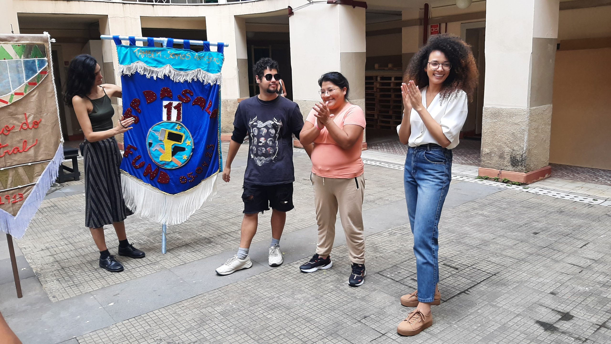 Foto colorida mostrando Marina Lima e três estudantes do curso de Técnicas de Palco. No lado esquerdo da foto uma das estudantes segura um estandarte na cor azul com o escrito "TP DA SALA 11. FUND 05.04.2023"
