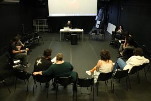 Foto colorida mostrando os integrantes do curso "O Lugar da Fotografia na Cena Teatral Paulistana" sentados em uma roda e Bob Sousa sentado na mesa compondo esse círculo