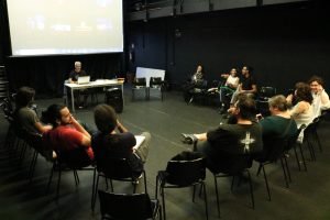 Foto colorida mostrando os integrantes do curso "O Lugar da Fotografia na Cena Teatral Paulistana" sentados em uma roda e Bob Sousa sentado na mesa compondo esse círculo