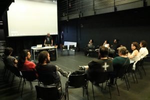 Foto colorida mostrando os integrantes do curso "O Lugar da Fotografia na Cena Teatral Paulistana" sentados em uma roda e Bob Sousa em pé apoiado na mesa compondo esse círculo