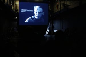 Foto colorida com luz escura mostrando uma imagem no telão da sala do curso de extensão cultural com Bob Sousa