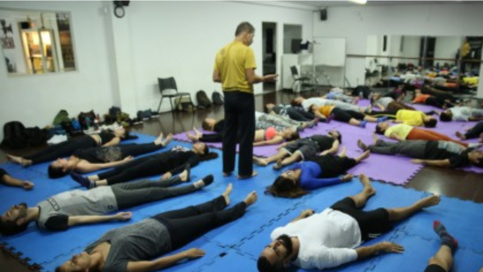 Foto colorida mostrando pessoas deitadas no tatame fazendo uma prática de Feldenkrais e o condutor no meio deles em pé