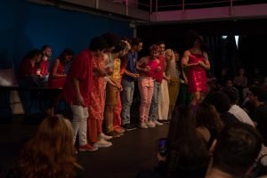 Fotografia colorida mostrando cena da apresentação de encerramento do curso de extensão cultural de Teatro Musical, com os integrantes de mãos dadas