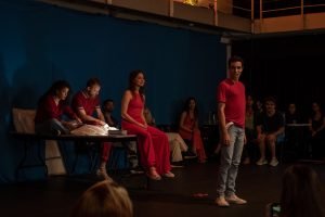 Fotografia colorida mostrando os integrantes do curso de extensão cultural de "Teatro Musical", com figurino na cor vermelha, em apresentação de encerramento