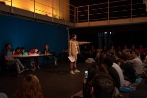 Fotografia colorida mostrando cena da apresentação de encerramento do curso de extensão cultural de Teatro, com um dos integrantes de figurino em tons pasteis à frente