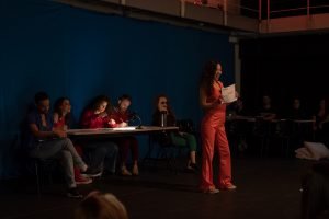 Fotografia colorida mostrando cena da apresentação de encerramento do curso de extensão cultural de Teatro, com uma das integrantes de figurino alaranjado à frente mostrando um papel branco