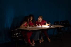 Fotografia colorida mostrando cena da apresentação de encerramento do curso de extensão cultural de Teatro. Na cena tem uma mesa sobre ela uma luminária e três integrantes sentados com figurino avermelhado