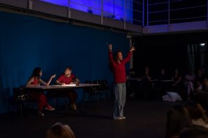 Fotografia colorida mostrando cena da apresentação de encerramento do curso de extensão cultural de Teatro, com um dos integrantes de figurino avermelhado à frente com os braços levantados