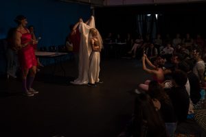 Fotografia colorida mostrando cena da apresentação de encerramento do curso de extensão cultural de Teatro. Na cena tem um tecido branco esticado e uma das integrantes na frente