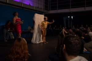 Fotografia colorida mostrando cena da apresentação de encerramento do curso de extensão cultural de Teatro. Na cena tem um tecido branco esticado e um dos integrantes na frente com a mão esquerda apontando para frente