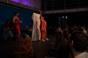 Fotografia colorida mostrando cena da apresentação de encerramento do curso de extensão cultural de Teatro. Na cena tem um tecido branco esticado e uma das integrantes na frente levantando um de seus braços