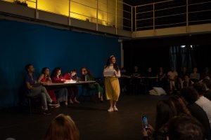 Fotografia colorida mostrando cena da apresentação de encerramento do curso de extensão cultural de Teatro, com um dos integrantes de figurino amarelado à frente segurando um papel branco