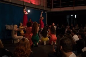 Fotografia colorida mostrando cena da apresentação de encerramento do curso de extensão cultural de Teatro, com os integrantes abaixados apontando para um deles que está de pé no centro e com uma de suas mãos levantadas