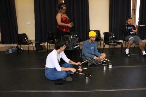 Fotografia colorida mostra estudantes e Tâmara tocando instrumentos criados por eles mesmo em aula com Barulho Max