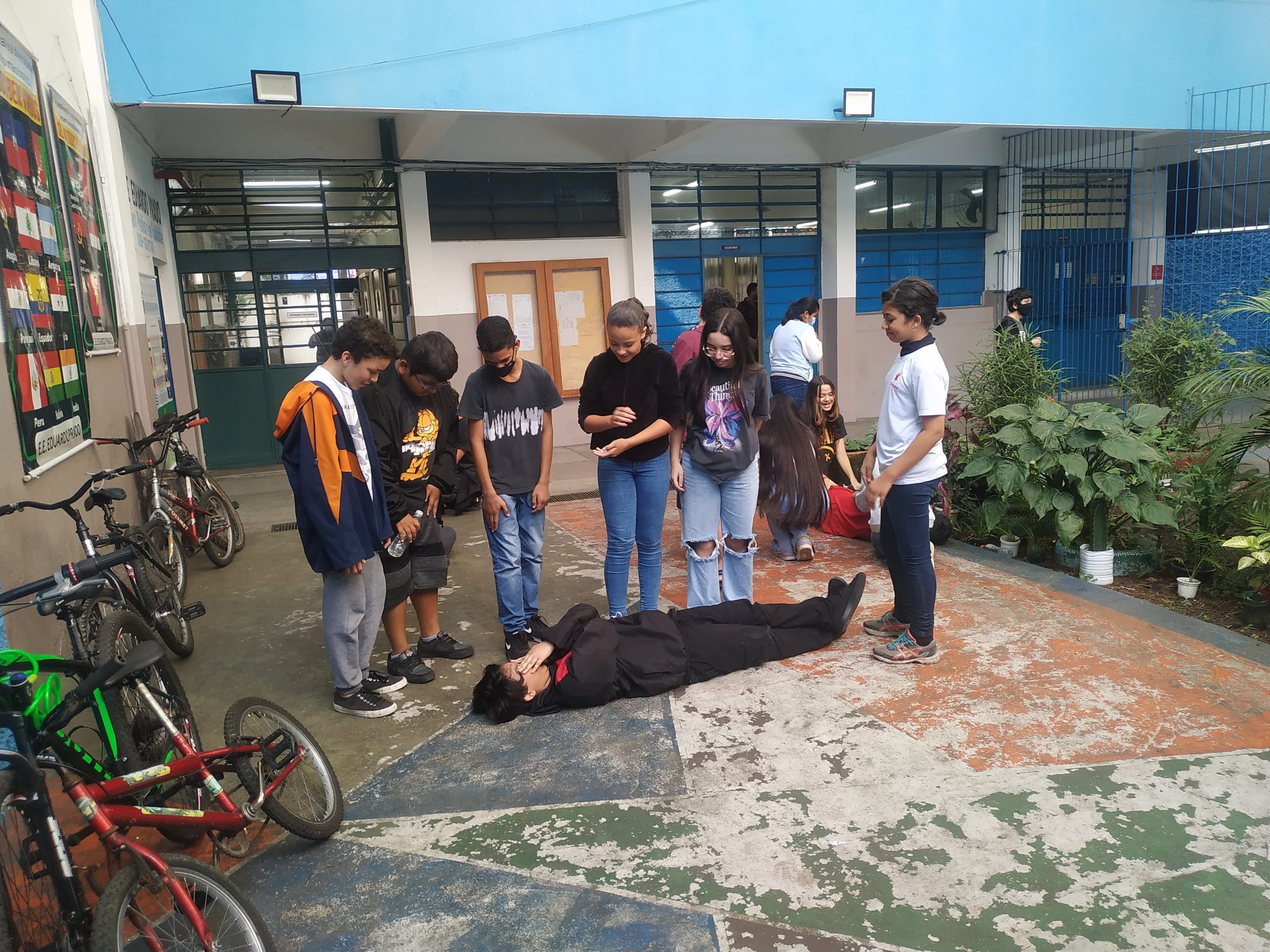 Fotografia colorida mostrando seis estudantes da E.E. Eduardo Prado em pé olhando para um outro estudante deitado no chão
