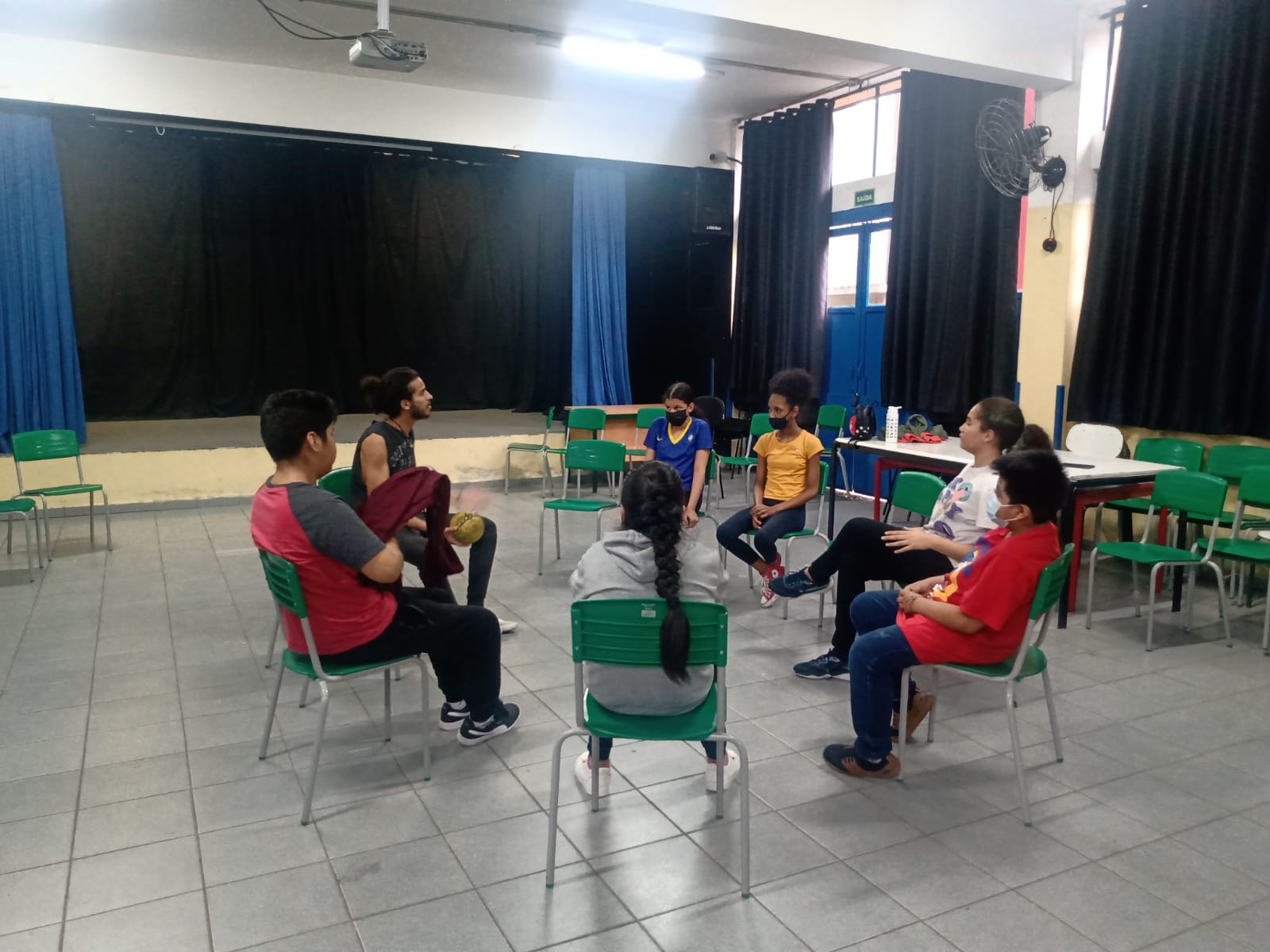 Fotografia colorida mostrando os participantes da oficina de jogos teatrais sentados em círculo, em uma das salas da E.E. Frei Paulo Luig