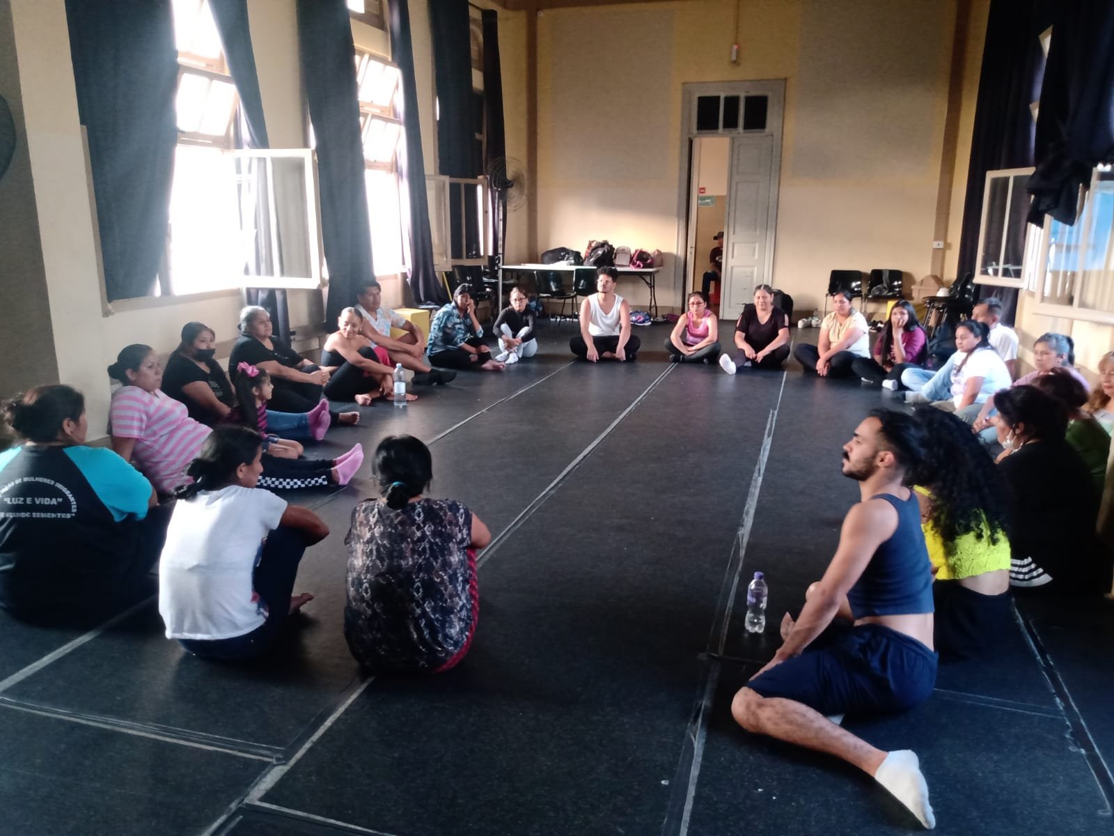 Fotografia colorida mostrando os participantes da oficina de técnicas de teatro sentados no chão em círculo, em uma das salas da sede Brás