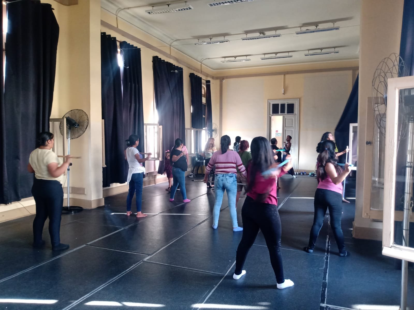 Fotografia colorida mostrando os participantes da oficina de técnicas de teatro segurando cabos de vassoura na horizontal, em uma das salas da sede Brás