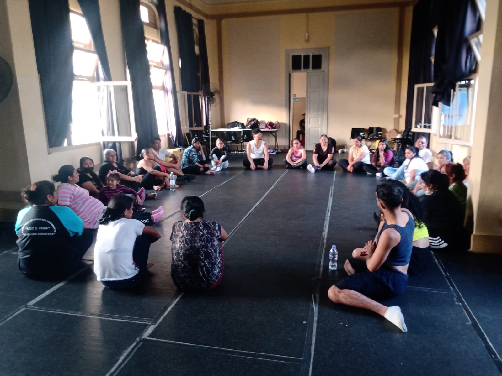 Fotografia colorida mostrando os participantes da oficina de técnicas de teatro sentados no chão em círculo, em uma das salas da sede Brás