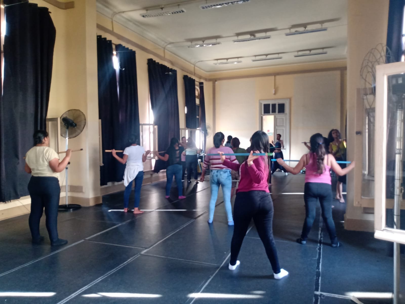 Fotografia colorida mostrando os participantes da oficina de técnicas de teatro segurando cabos de vassoura na horizontal, em uma das salas da sede Brás