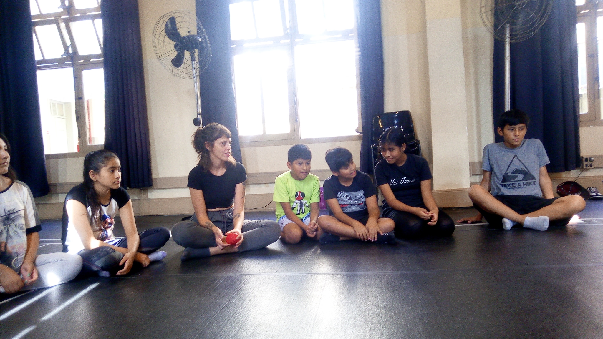 Fotografia colorida mostrando cinco crianças e a estudante da SP sentadas no chão na oficina de jogos teatrais realizadas na sede Brás em parceria com a Amilv