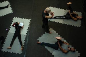 Fotografia colorida mostra aula do curso de "Afinação do Corpo". Três participantes estão deitados nos tatames individuais e o orientador em pé.