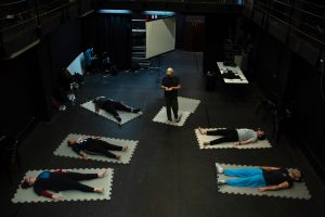 Fotografia colorida mostra aula do curso de "Afinação do Corpo". Os participantes estão deitados em tatames individuais e o orientador em pé.
