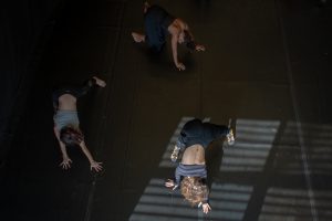 Fotografia colorida mostra aula do curso de "Dança Contemporânea". Três participantes fazem movimentos no chão