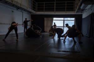 Fotografia colorida mostra aula do curso de "Dança Contemporânea". Todos estão fazendo movimentos diversos no chão