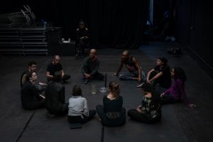 Fotografia colorida mostra aula do curso de "D´água movimento do corpo". O orientador e os participantes estão sentados no chão formando um círculo