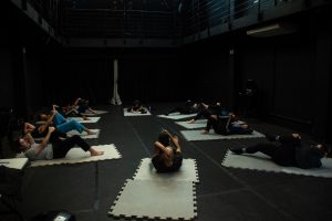 Fotografia colorida mostra aula do curso de "Afinação do Corpo". Todos estão deitados em tatames individuais em prática de alongamento.