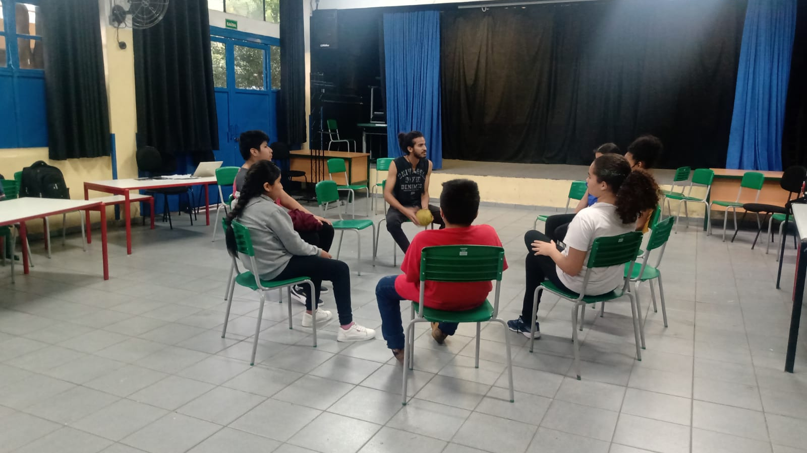 Fotografia colorida mostrando os participantes da oficina de jogos teatrais sentados em círculo, em uma das salas da E.E. Frei Paulo Luig 